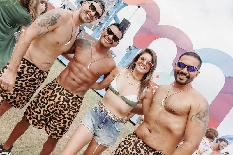 Três homens sem camisa, de bermuda estampada de onça, abraçados a uma mulher de biquíni, sorrindo diante do palco azul