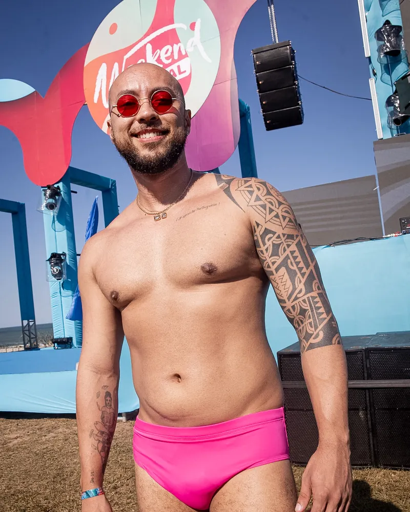 Homem de sunga pink e óculos vermelhos, de pé, com o palco azul e o mar atrás