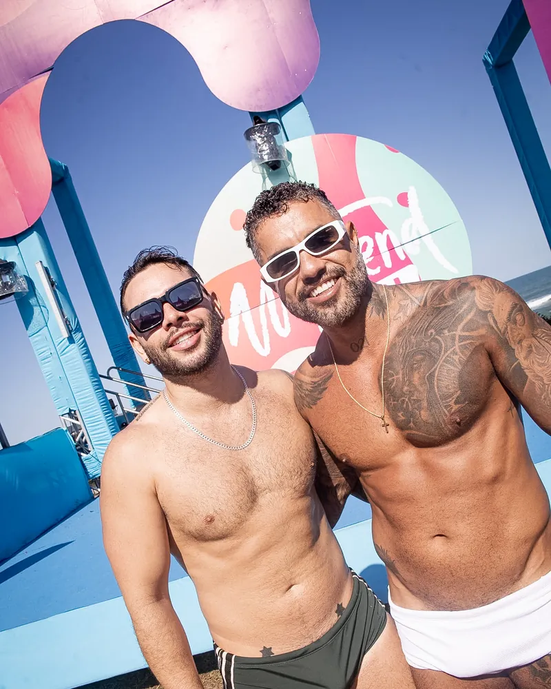 Dois homens sem camisa, de sunga e short, abraçados e sorrindo diante do logo do Weekend Festival