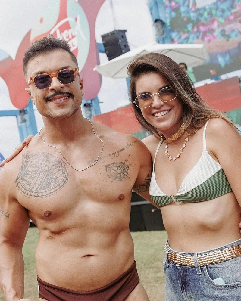 Homem de sunga marrom com tatuagens no peito, sorrindo, ao lado de uma mulher de biquíni