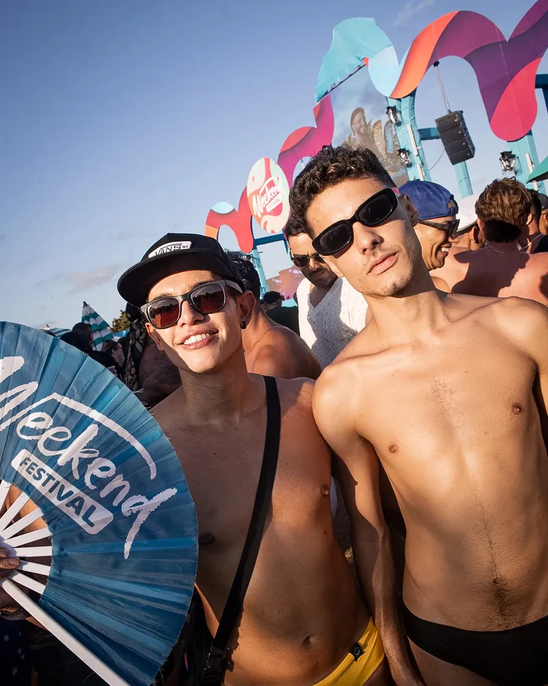 Dois homens sem camisa, um de sunga amarela, sorrindo ao lado de um leque do Weekend Festival